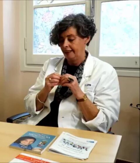 Medico donna con camice bianco seduta alla scrivania durante un consulto, con opuscoli informativi e un libro su menopausa e salute della donna.